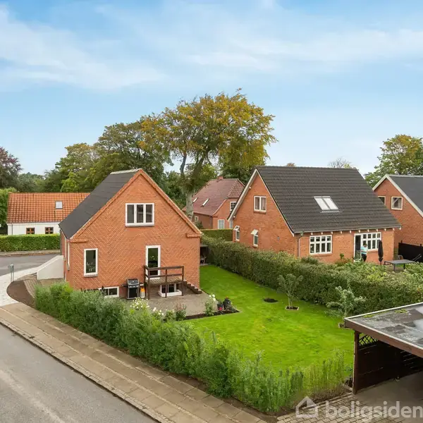Rødt murstenshus med have, stillestående, i et villakvarter med græsplæne og carport. Flere huse og træer omgiver stedet, under en blå himmel.