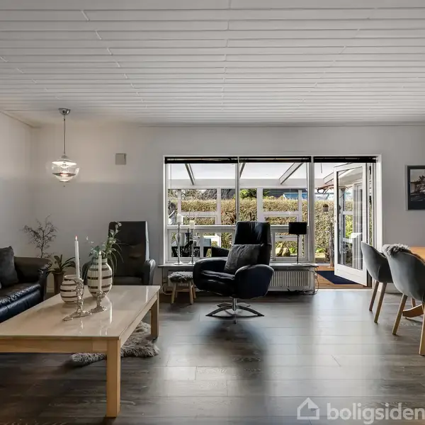 Stue med sort lædersofa og sofabord med dekorationer, ved siden af spisebord med grå stole. Glasdør åbner til overdækket terrasse med udsigt til have.
