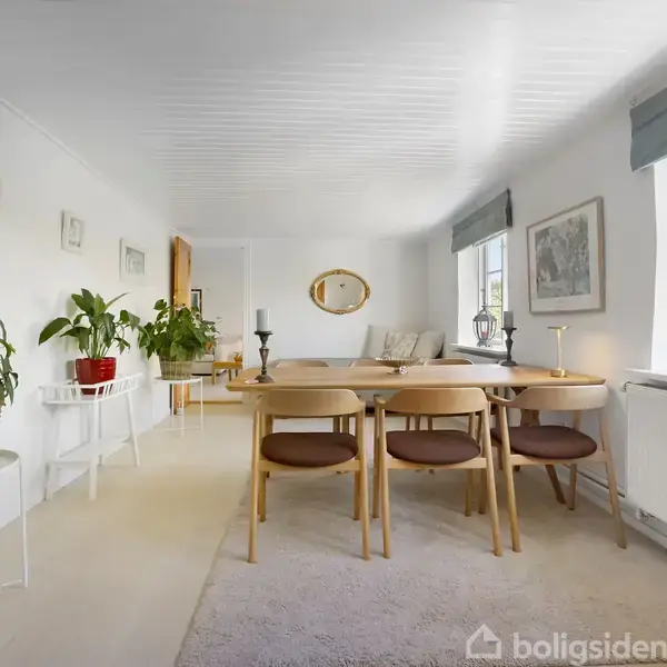 Et spisebord med stole står i en lys stue. Bordet er dekoreret med lysestager. Store vinduer til højre slipper dagslys ind. Planter pynter langs væggen.