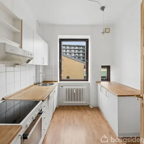 Et lyst køkken med træbordplader og hvide skabe. En ovn og kogeplade er installeret. Udsigt gennem vinduet viser en bygning. Radiator under vinduet, trædør til højre.