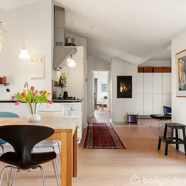 Et lyst og moderne køkken-alrum med spisebord og farverige stole. En køkkenø med blomster står forrest. En gang med tæppe fører til et lyst rum med kunst på væggen.