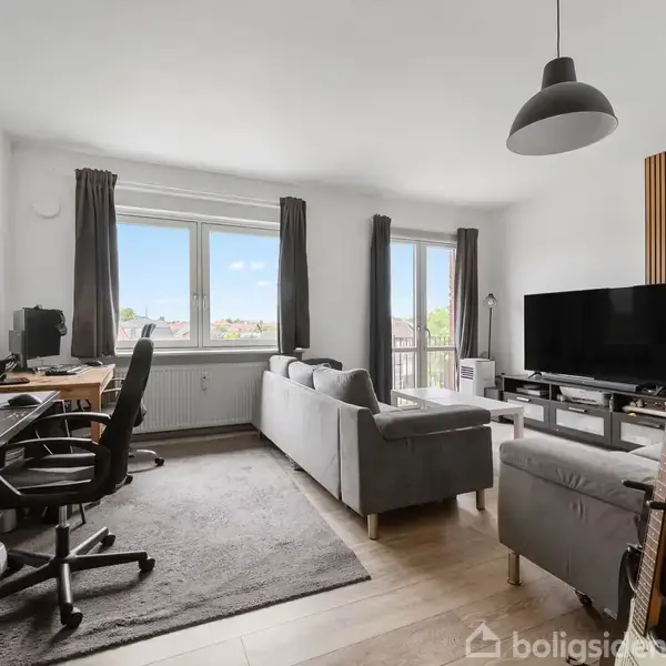 En stue med en grå sofa arrangeret i en L-form, et skrivebord med computer, en fladskærms-tv på reol. Trægulve og store vinduer med gardiner giver naturligt lys.