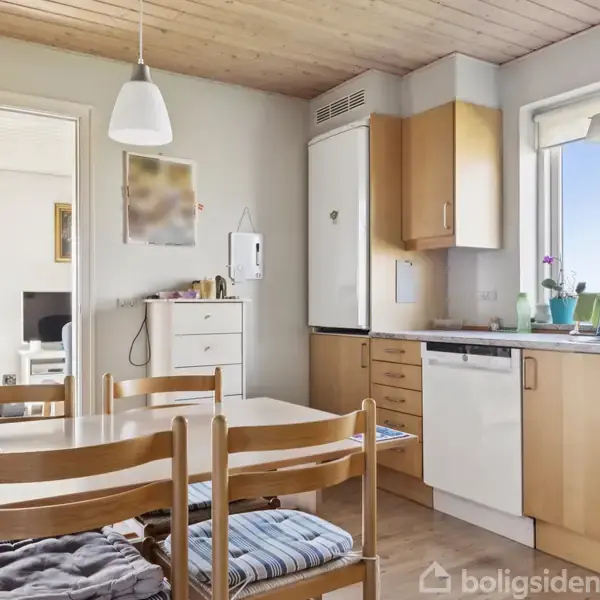 Køkkenbord med stole står samlet i et lyst køkken med træskabe. En stor vindue lader lys ind og viser udsigt over landskab. Der er også en opvaskemaskine og hængende lamper.