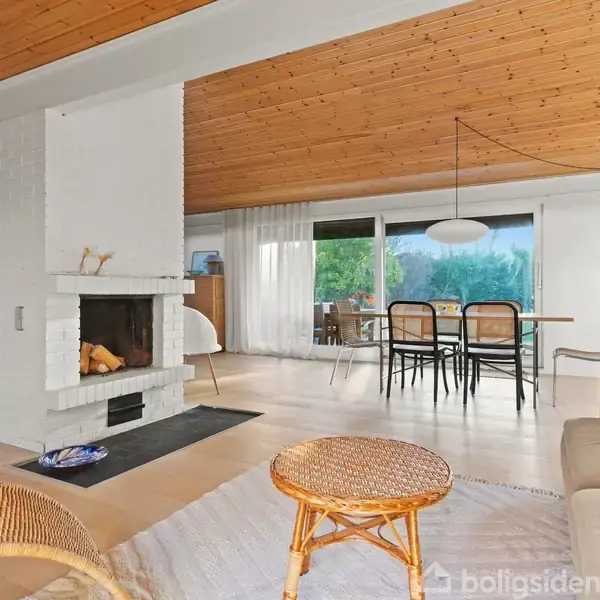 Brændeovn i stue opvarmer rummet, mens en spisebordsgruppe står ved store vinduer. Trægulv og træloft skaber en varm og indbydende atmosfære. Sofaer og kurvemøbler tilføjer komfort.