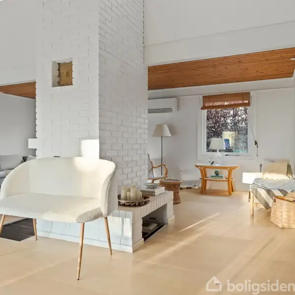 En hvid sofa står foran en hvidmalet pejs i et lyst rum med trægulve. Store vinduer lader sollys ind, og der er moderne møblering med en kurv i hjørnet.