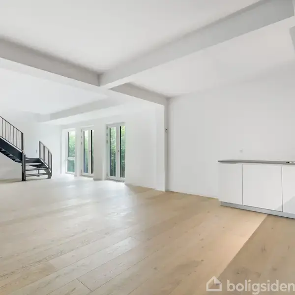 En rummelig, lys stue med trægulv og en minimalistisk hvid køkkensektion til højre. Trappe fører op til en etage, og store vinduer lukker naturligt lys ind i rummet.