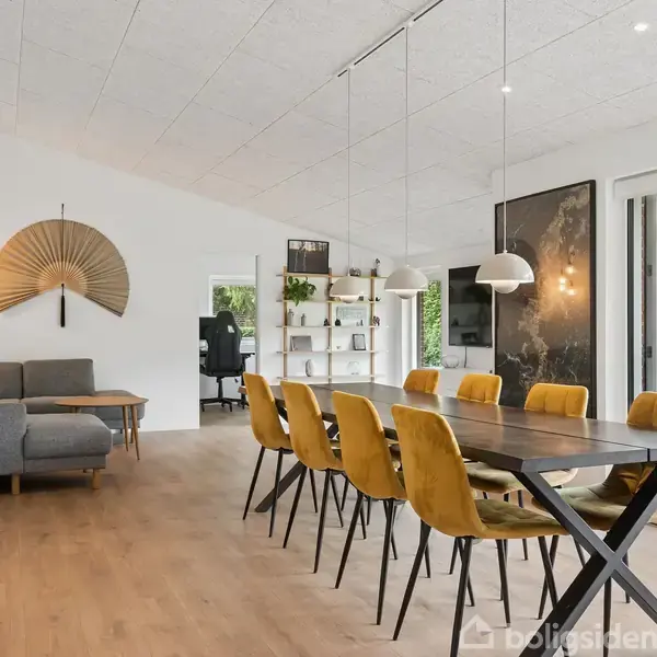 En spisebord med gule stole står i en moderne stue. Ved siden af er en grå sofa og dekorative elementer på væggen. Store vinduer viser en have udenfor.