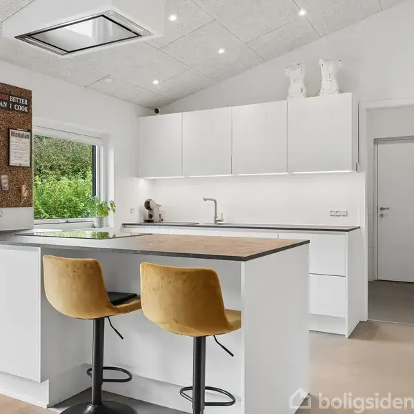 Et moderne køkken med hvide skabe og en sort bordplade. To gule barstole står ved øen. Vindue til venstre, opslagstavle med noter. Lysegrå gulv og loft.