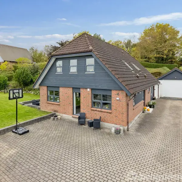 Et murstenshus med mørkt tag og skodder står midt i en gårdsplads med fliser, omgivet af grønne haver. Til højre ses en indkørsel, der fører til en garage. En basketballkurv står på græsplænen.