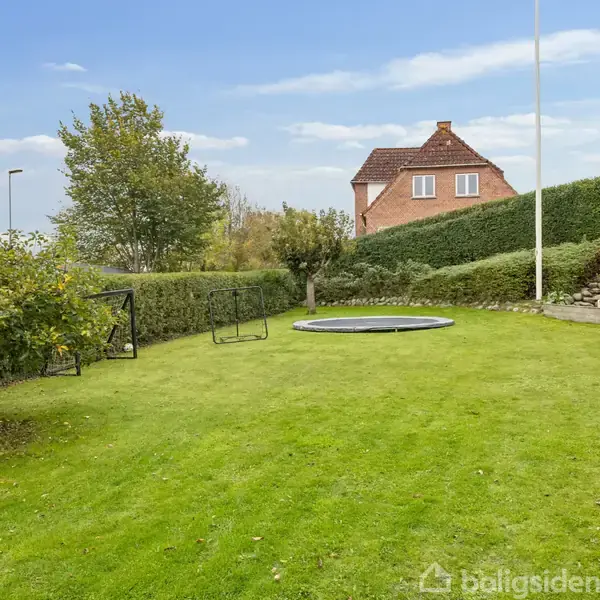 Græsplæne med nedgravet trampolin omgivet af hække og træer. I baggrunden ses et murstenshus med rød tagsten.