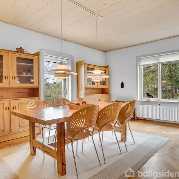 Et træbord omgivet af seks stole står i en spisestue. To lamper hænger over bordet. Store træskabe står op ad væggen. Et vindue viser grønne træer udenfor.