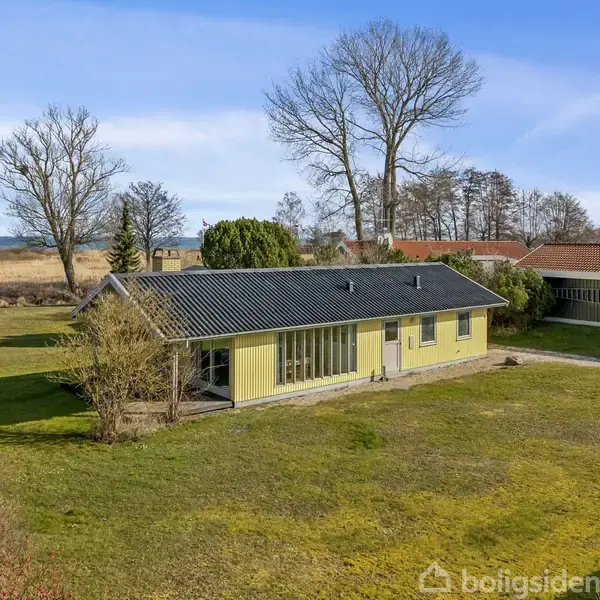 Et gult hus med sort tag står på en grøn græsplæne. Det er omgivet af træer, med havudsigt i baggrunden under en blå himmel.