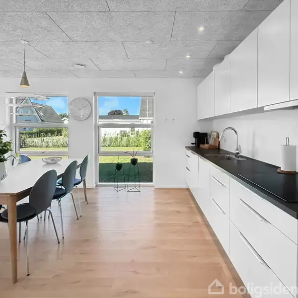 Køkkenbord med sorte overflader og hvide skabe; ved siden af spisebord med stole. To vinduer giver udsigt til en have. Fjernsyn og vægur er på væggen.