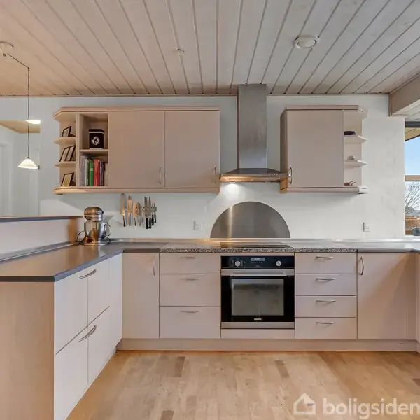 Et moderne køkken med hvide skabe og sorte bordplader. Ovn og emhætte midt i billedet. Store vinduer i højre side giver naturligt lys. Knive hænger på væggen.