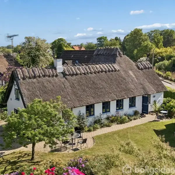 Et traditionelt bindingsværkshus med stråtag står omgivet af en frodig have. Huset har hvid facade og blå skodder. En terrasse med havemøbler er synlig til venstre; omgivelserne er grønne.