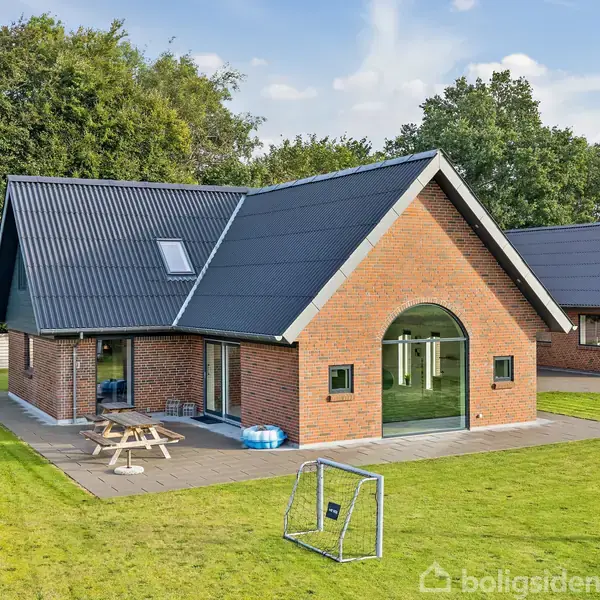 Et rødstenshus har et mørkt skråtag, placeret i en grøn have med legeudstyr og et picnicbord. Omringet af græsplæne samt træer i baggrunden.