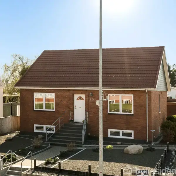 Et rødt murstenshus står stille i en forstadskontekst med træer og veje omkring. Huset har en enkelt indgangstrappe og dør. Tekst på skilt: "Til salg - 75 52 71 11."