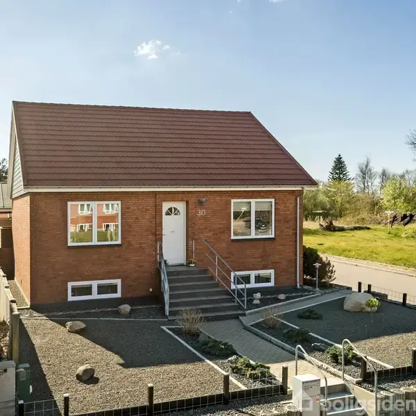 Et rødt murstenshus med trefagsvinuer står i en forhave med grus og små buske. En trappe fører op til døren, og nummeret "30" er ved siden af.