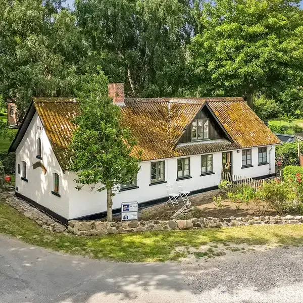 Et hvidt, ældre hus med et mosgroet tegltag er omgivet af grønne træer og en grusvej. En ejendomsmægler skilt ses i forhaven.