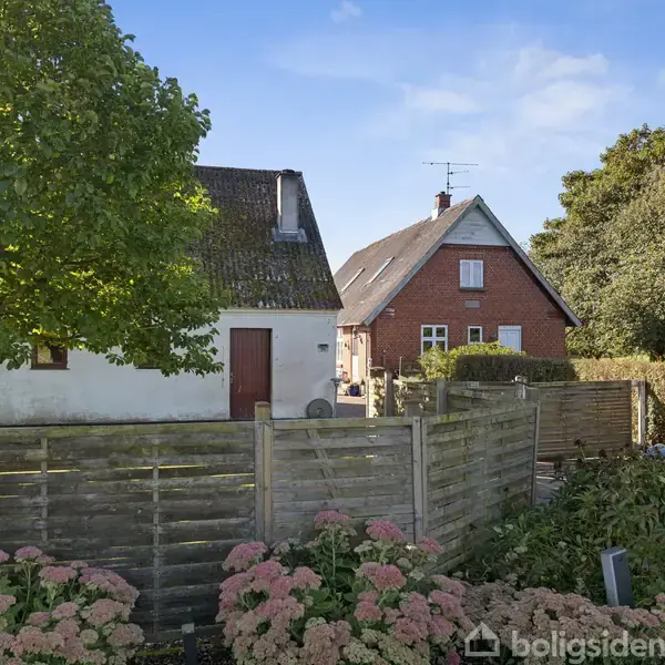 Et rødt og hvidt hus står bag en træhegn. En stor grøn træ til venstre. Omkringliggende have med blomster og buske. Solskin og blå himmel i baggrunden.