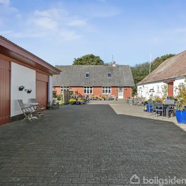 En gårdsplads omgivet af tre bygninger; midten er et rødt hus med vinduer, flankeret af to hvide bygninger. Der er blomster i krukker og udemøbler rundt omkring.