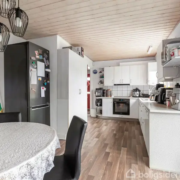 Et køkken med et rundt spisebord dækket med dug og frugtskål, køleskab med magneter i hjørnet, og hvide skabe langs væggen. Rummet har lyst trægulv og loft.