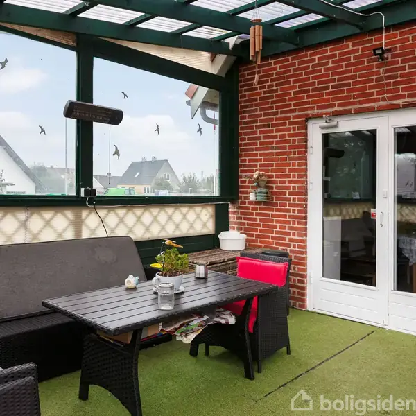 Terrasse med sofa og bord arrangeret på kunstgræs. Røde murstensvægge og vinduer med fugledekorationer danner rammen. To glasdøre fører ud mod det udendørs område.