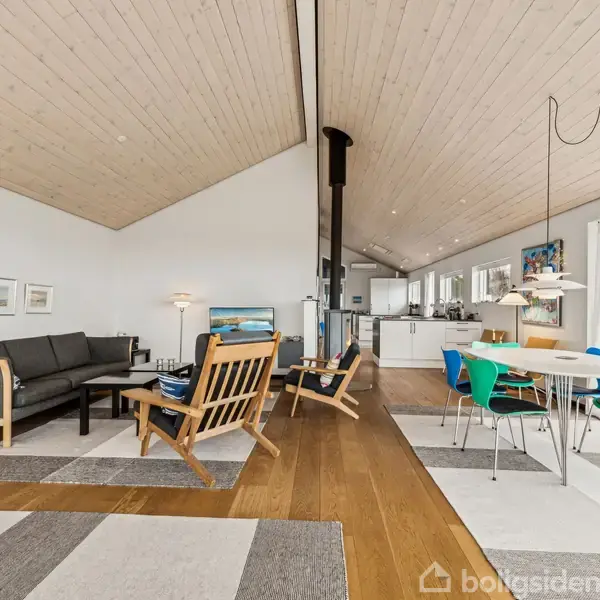 Pejs opvarmer moderne stue i et hus med store vinduer, der afslører en naturudsigt. Træmøbler og grønne stole omkranser et spisebord under loftslampe. Ingen synlig tekst.