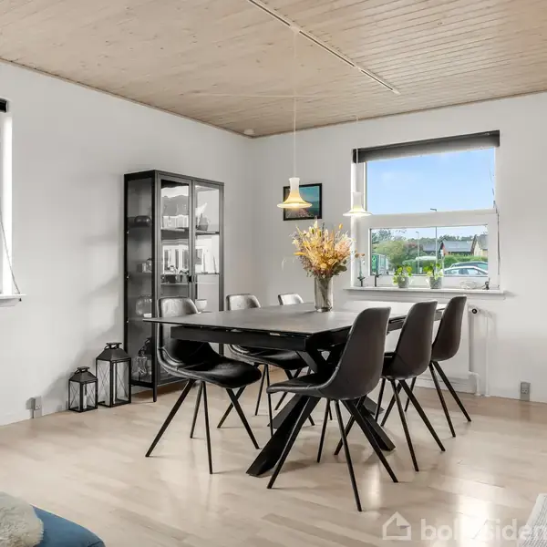 Et spisebord med seks sorte stole står i en lys spisestue med trægulv. Omkring er der hvide vægge, vinduer med udsigt til træer, og en reol med dekorationer.