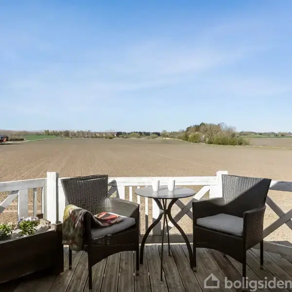 To kurvestole og et rundt bord står på en terrasse. En vid udsigt over en flad mark og blå himmel danner baggrunden.