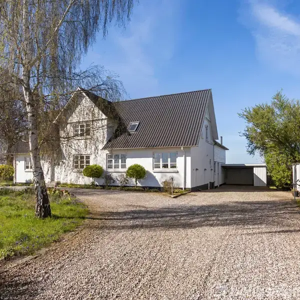 Hvidt hus står roligt med grusvej foran, omgivet af træer og en blå himmel. En garage er ved siden af huset til højre.