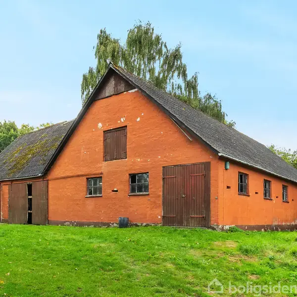 En stor, orange lade står stille på en grøn græsplæne, omgivet af frodig vegetation og træer, under en klar blå himmel.