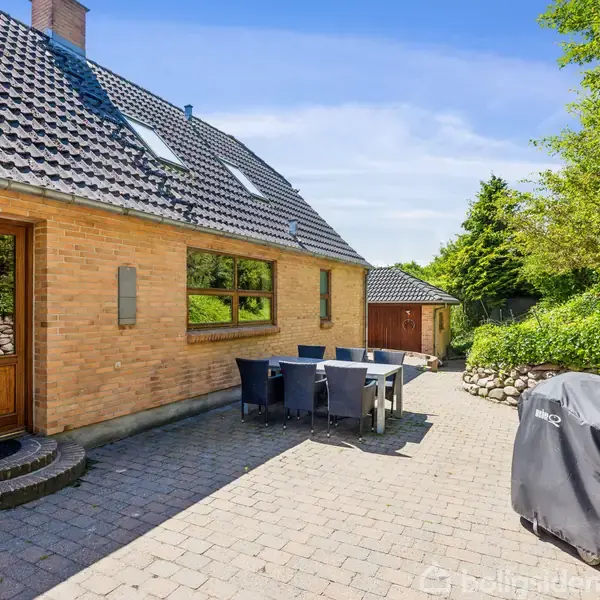 Et murstenshus med skråt tag har en flisebelagt terrasse med spisebord og stole. Ved siden af står en grill dækket med et betræk. Omgivelserne består af grønne buske og træer.