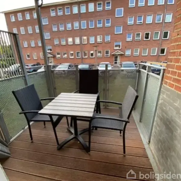 To stole og et bord står på en altan med trægulv, omkranset af gennemsigtige glasvægge. I baggrunden ses en rød murstensbygning med parkering foran.
