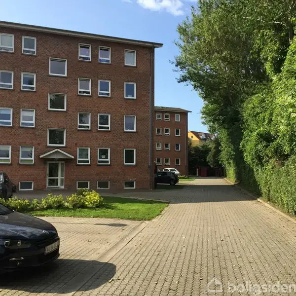 Et rødt murstensbyggeri står stille i en stille gårdsplads med parkerede biler og omgivet af grøn beplantning og hække.
