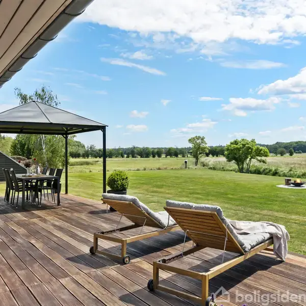 Terrasse med to liggestole og et spisebord under en pavillon. Græsplæne og træer strækker sig ud mod en åben mark under en blå himmel med hvide skyer.