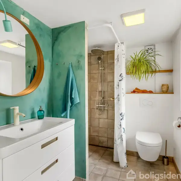Hvid håndvask med rund spejl, handlingsløs, i et lyst badeværelse med grøn væg, bruser med forhæng, og toilet ved siden af en hylde med plante. En blå håndklæde hænger ved spejlet.