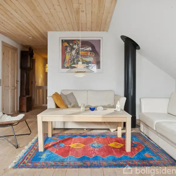En stue med to hvide sofaer står omkring et træbord på et farverigt tæppe. En væg har moderne kunst, og rummet har et skråloft og træpaneler.