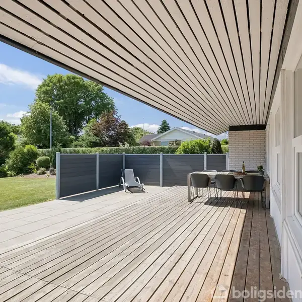 Træterrasse strækker sig langs en husvæg med havemøbler, inklusiv et bord og stole. Omkranset af en græsplæne og træer, skabt til udendørs afslapning.