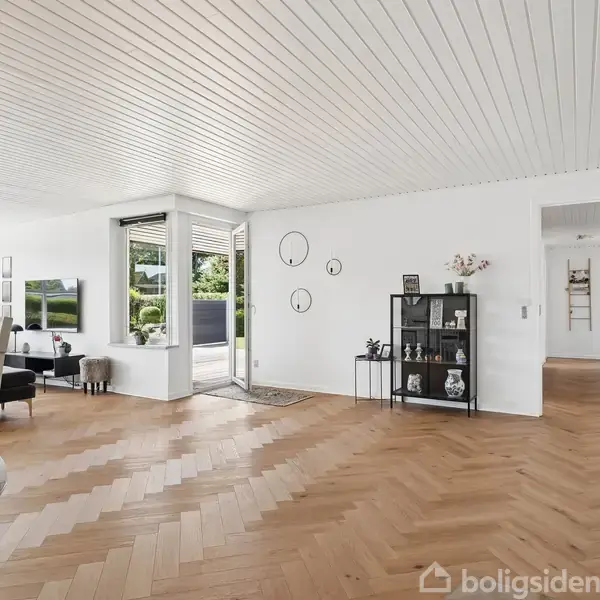 Stue med trægulv og brændeovn; hygge skabes i et lyst rum med hvidt loft og vægge. Møbler inkluderer sofa, reol, samt dekorative ure på væggen. Udsigt til have.