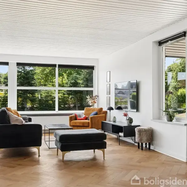 En moderne stue med sorte sofaer og en gul stol foran et stort vindue. En fladskærms-tv hænger på væggen. Udenfor ses en terrasse og grønne træer.