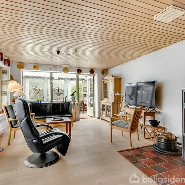 Spisestue med træbord og stole, set i hjem med trægulv og loft. Et stort vindue viser natur udenfor. Til venstre ses en lænestol og reoler med dekorative genstande.