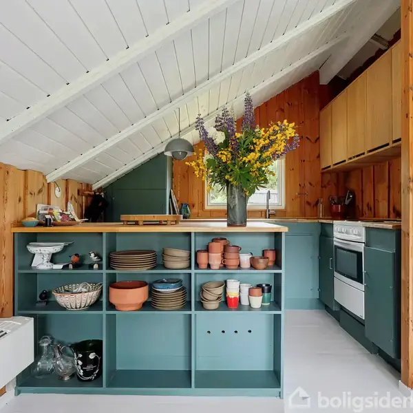 Køkken med åbne hylder indeholder tallerkener, skåle og kopper. Stor vase med gule og lilla blomster står på bordpladen. Trævægge og skrånende hvidt loft skaber en hyggelig atmosfære.