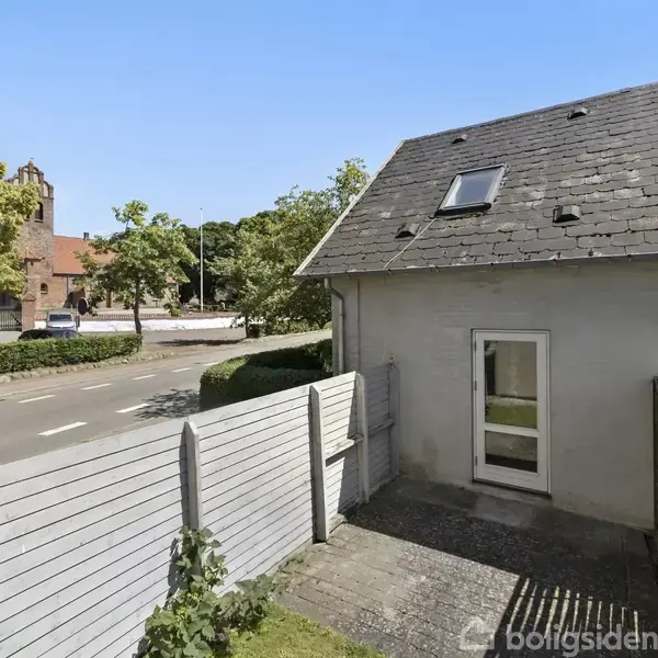 Et gråt, skråt hus med et vindue og en dør står ved siden af en vej. En hvid træhegn afspærrer området foran huset. I baggrunden ses grønne træer og en rød murstensbygning.