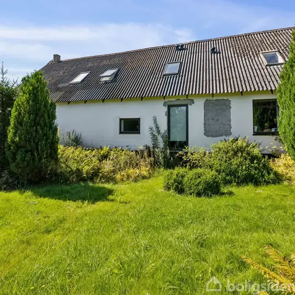 Et hus med hvid facade og skråt tag har flere ovenlysvinduer. Det står i en grøn have omgivet af buske og træer under en klar blå himmel.