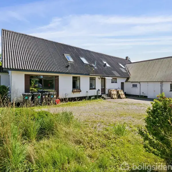 En hvid bygning med sort skråtag står stille i en frodig have. Gruset gårdsplads foran og grønne buske omgiver huset.
