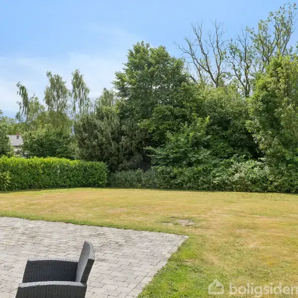 Terrassemøbler står på en flisebelagt terrasse; græsplænen breder sig ud omkring det. Tæt bevoksning og træer markerer baggrunden, hvilket skaber en privat og grøn udsigt.