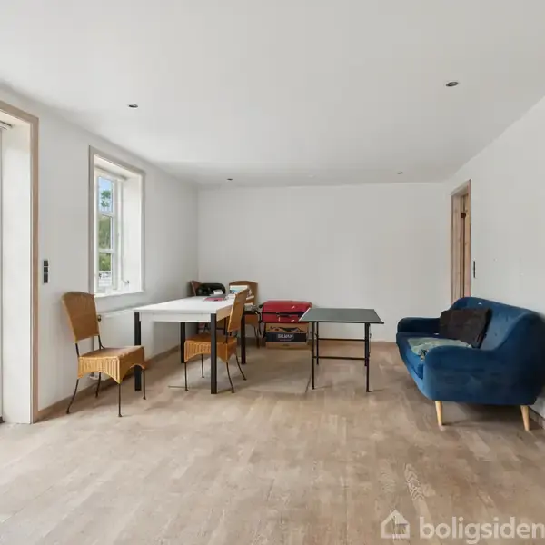 Et rum med parketgulv har et blåt sofaarrangement og et bord med stole. Store vinduer og glasdøre lukker dagslys ind, omgivet af en lys hvidmalet væg.