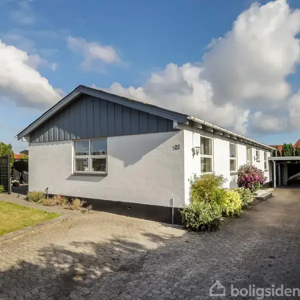 Et hvidt hus med blå gavl står omringet af en velplejet have. Tallet '28' er synligt på facaden. Flisebelagt indkørsel fører til en carport.