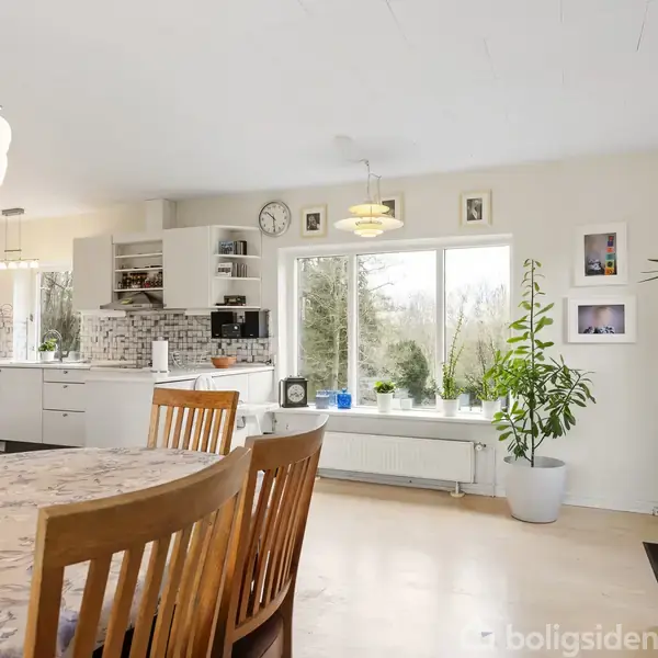 Et spisebord med blomsterdug, omringet af træstole, står i et lyst køkken. Store vinduer med udsigt til naturen dominerer væggen, omgivet af planter og moderne indretning.